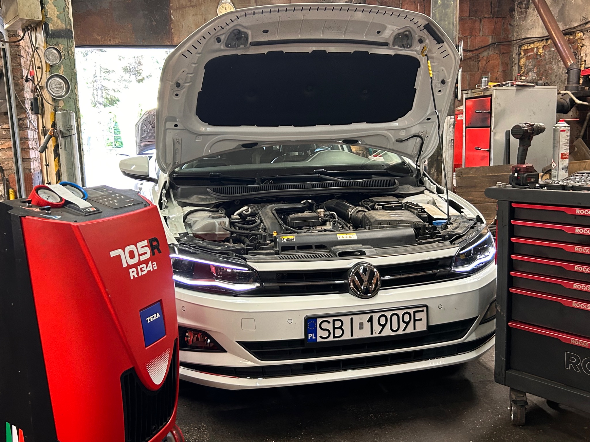 Biały samochód z otwartą maską w warsztacie, widoczna maszyna do klimatyzacji TEXA 705R R134a i wózek narzędziowy ROCKS. Tablica rejestracyjna SBI 1909F.