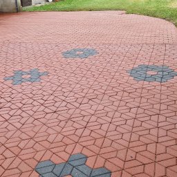 MS Serwis - Mycie Kostki Brukowej i Elewacji - Czerwona kostka brukowa ułożona we wzór z szarymi akcentami w kształcie kwiatów, prowadząca od bramy garażowej przez trawnik, po świeżym czyszczeniu.