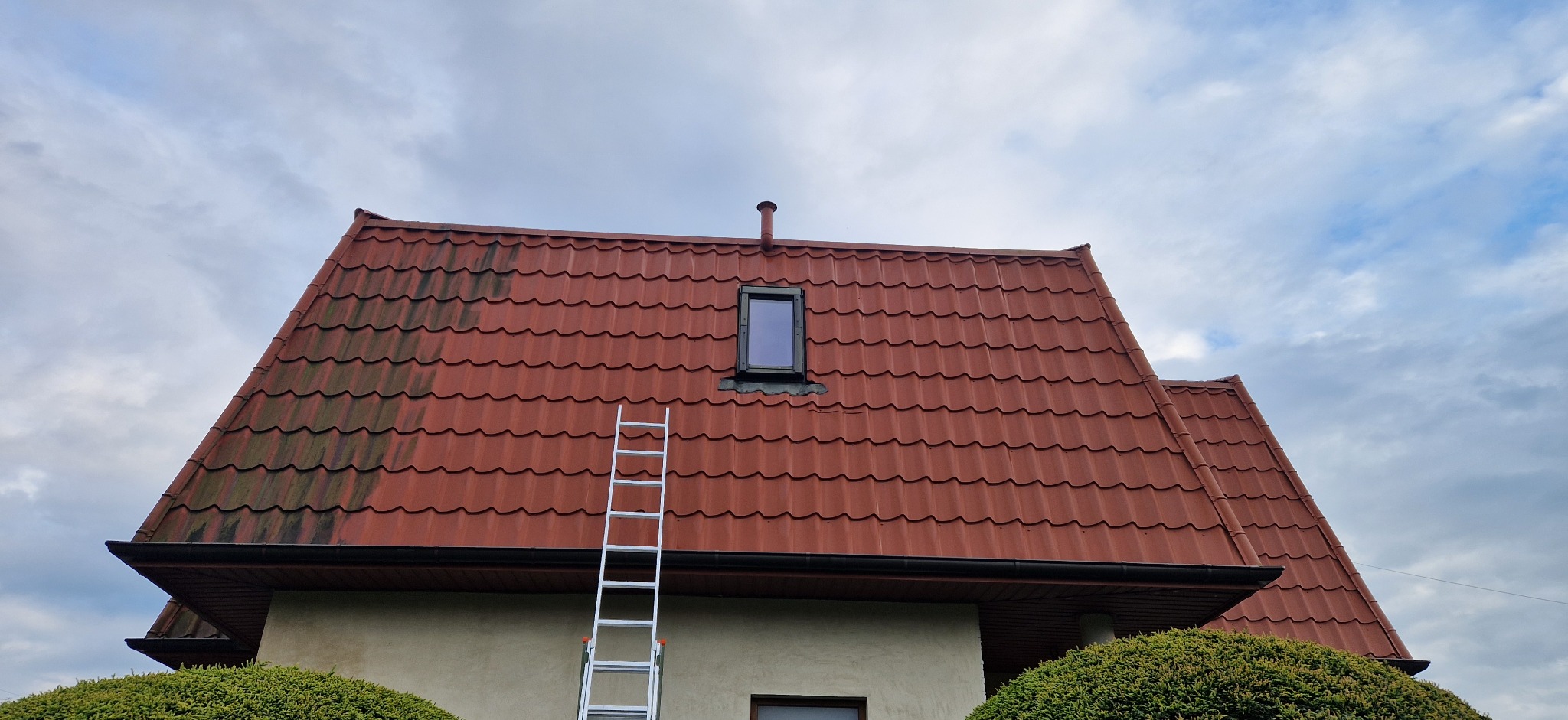 Częściowo umyty dach z czerwonej dachówki, widoczna drabina oparta o budynek, kontrast między czystą a zabrudzoną powierzchnią dachu, okno dachowe.