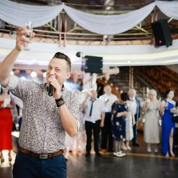 PRESTO - zespół na wesele - Uśmiechnięty konferansjer z mikrofonem i kieliszkiem na tle gości weselnych i nagłośnienia. Dekoracje sali bankietowej z białymi draperiami.