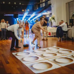 Goście weselni grają w kółko i krzyżyk na dużej macie z talerzami. W tle widoczna scena z oświetleniem i siedzący goście przy stołach. Sala balowa z parkietem.