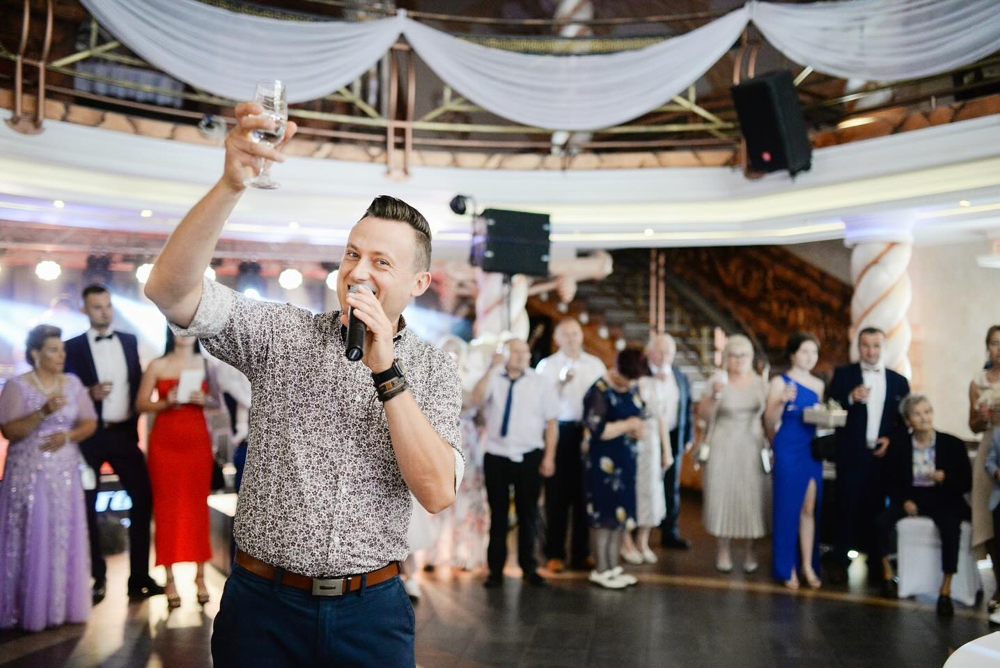 Uśmiechnięty konferansjer z mikrofonem i kieliszkiem na tle gości weselnych i nagłośnienia. Dekoracje sali bankietowej z białymi draperiami.