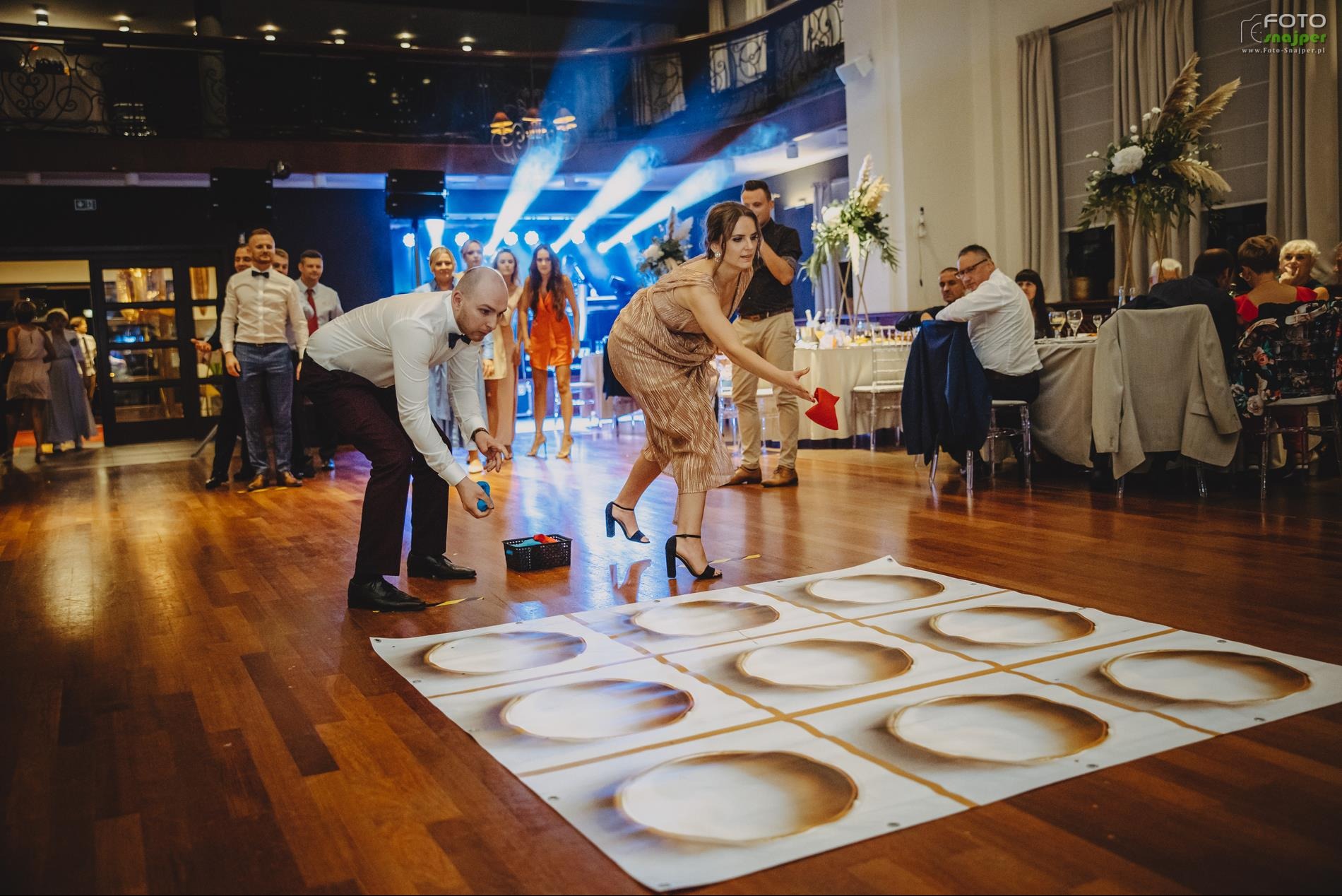 Goście weselni grają w kółko i krzyżyk na dużej macie z talerzami. W tle widoczna scena z oświetleniem i siedzący goście przy stołach. Sala balowa z parkietem.