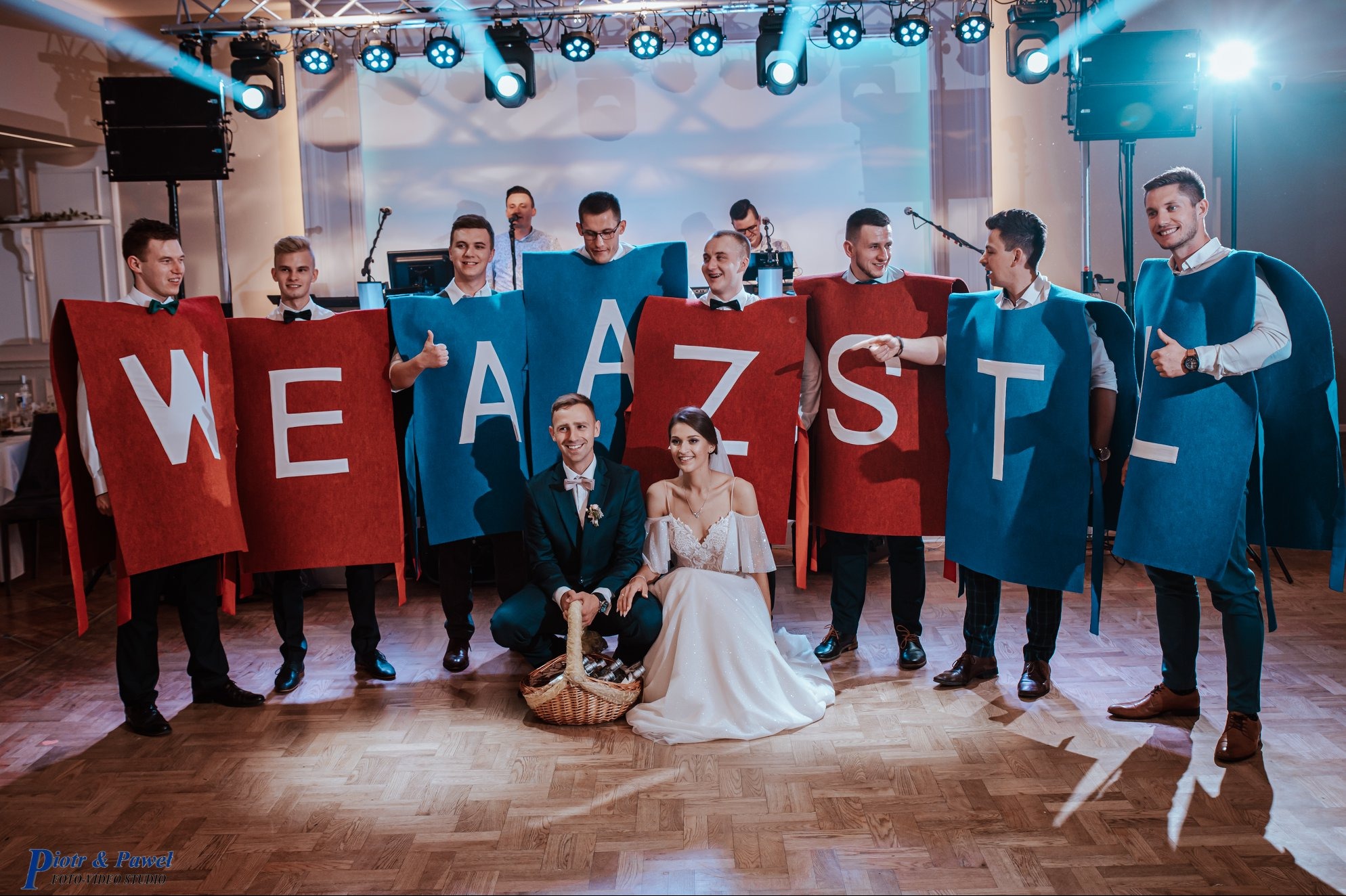 Zespół w kostiumach z literami 'WE AZ ST', para młoda klęczy z koszem na parkiecie, oświetlenie sceniczne, uroczysta atmosfera.