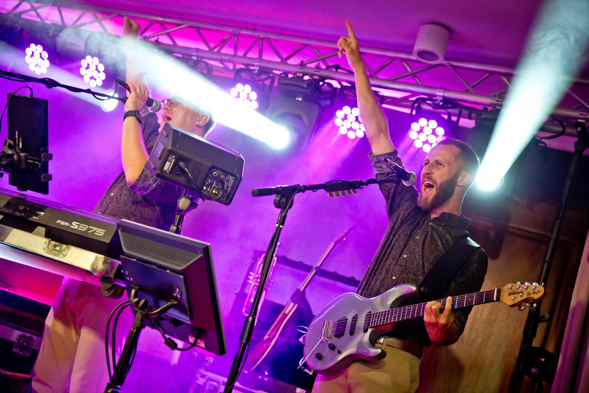 Zespół muzyczny w akcji: gitarzysta z brodą i klawiszowiec w okularach, unoszą ręce w górę, oświetleni fioletowymi światłami podczas występu na żywo.