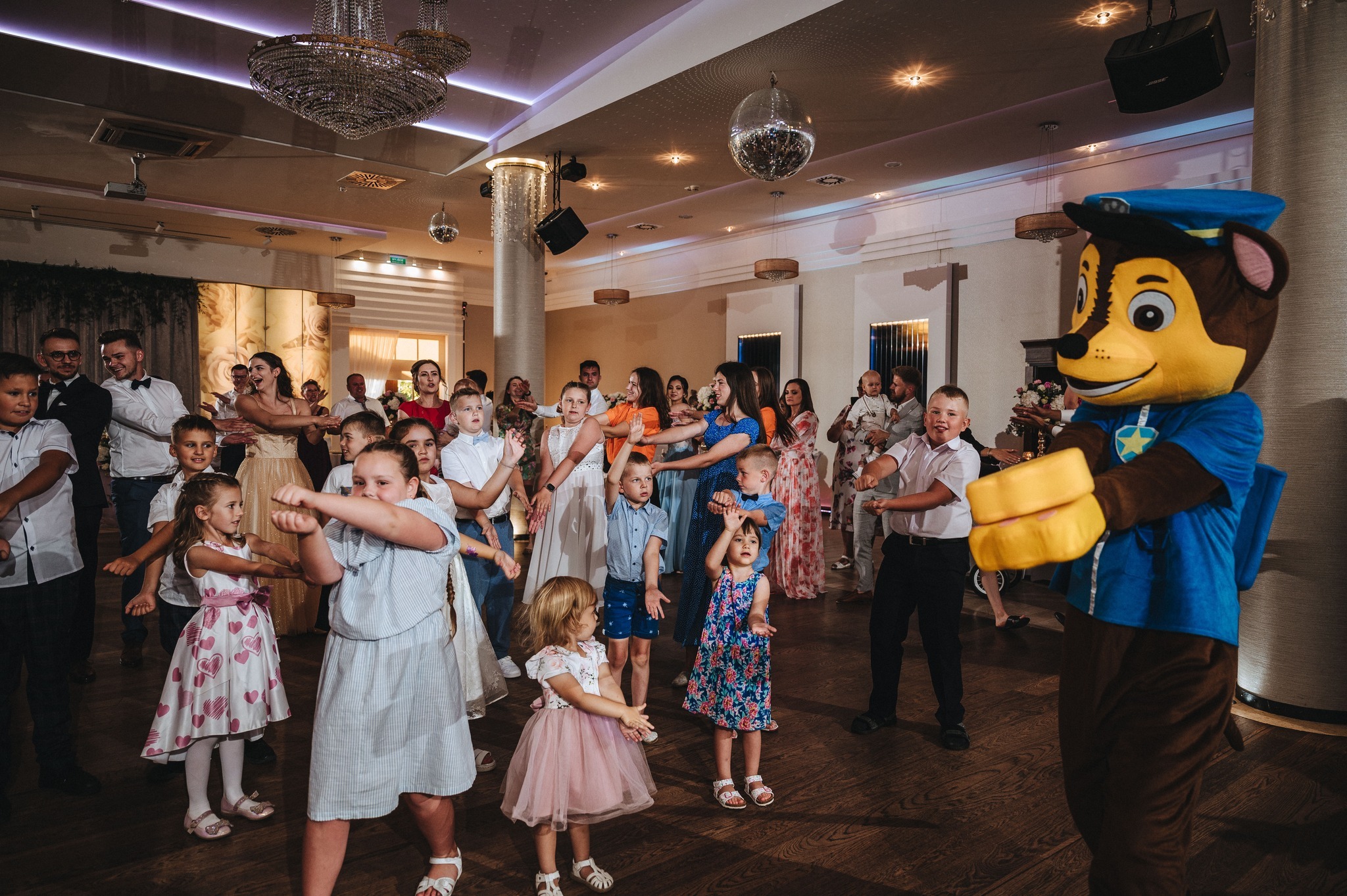 Animator w stroju Chase'a z Psiego Patrolu tańczy z dziećmi na sali balowej. W tle parkiet, lustrzana kula i żyrandole.