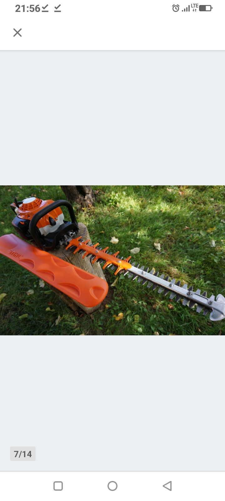 Pomarańczowo-srebrne nożyce spalinowe Stihl na pniu drzewa, obok osłona ostrza. Widoczne trawa i liście. Narzędzie do przycinania żywopłotu w ogrodzie.