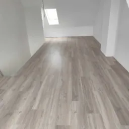 Jasne panele podłogowe w odcieniach szarości, ułożone w nowym pokoju na poddaszu z oknem dachowym i białymi ścianami. Widoczna klapa na suficie.