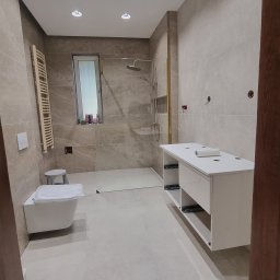 Complex - Jasna łazienka z beżowymi płytkami imitującymi kamień. Kabina prysznicowa ze złotymi detalami, biała szafka pod umywalkę i wisząca toaleta. Oświetlenie punktowe.