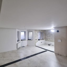 Complex - Jasne wnętrze w trakcie prac wykończeniowych: świeżo położona podłoga z płytek, częściowo pomalowane ściany, okna zabezpieczone folią malarską.