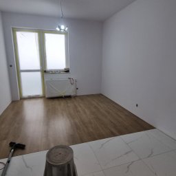 Complex - Układanie podłogi w Oławie: panele drewnopodobne zestawione z białymi płytkami. Okno zabezpieczone folią, grzejnik zakryty. Widoczne narzędzia.