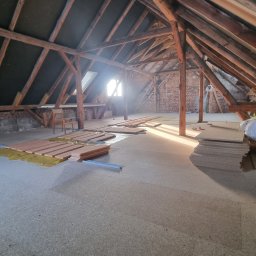 Complex - Adaptacja poddasza: drewniana konstrukcja dachu, izolacja podłogi, przygotowane panele podłogowe, ceglane ściany. W tle okno dachowe wpuszczające światło.