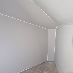 Complex - Szare wnętrze po malowaniu z widocznymi białymi liniami oddzielającymi ściany od sufitu i podłogi. Na podłodze widoczne ślady farby.