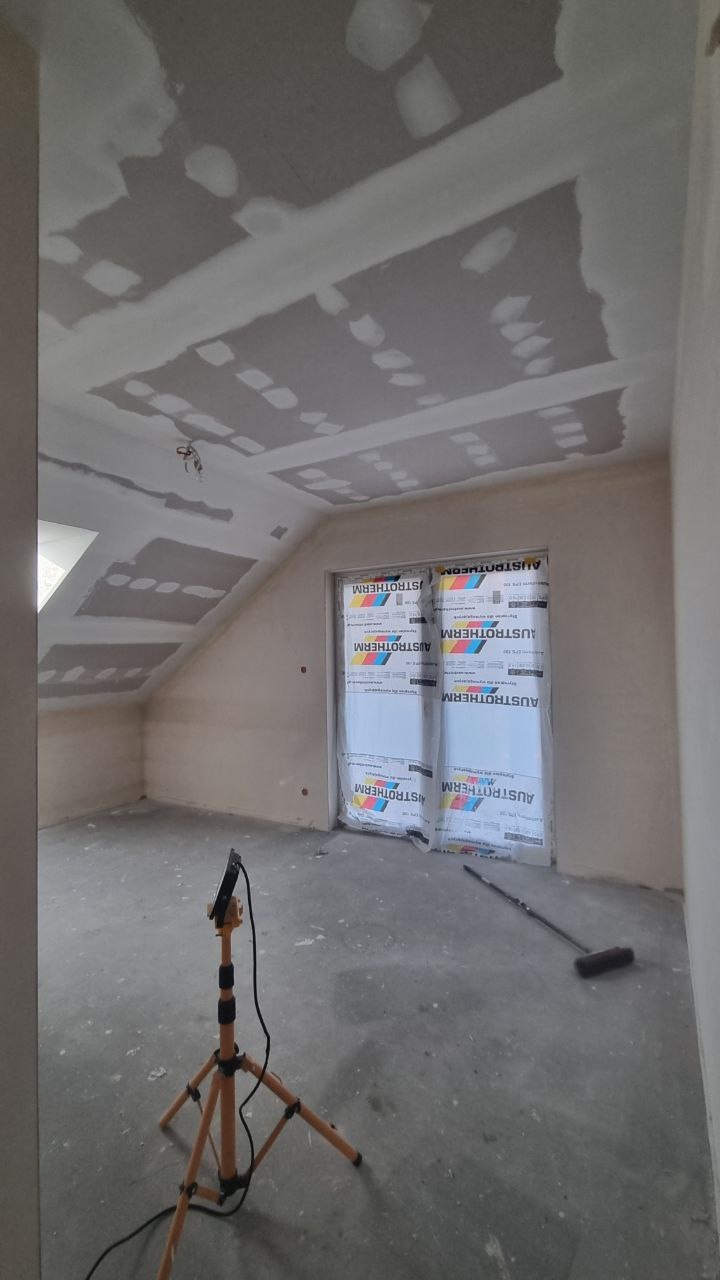 Pokój na poddaszu w trakcie prac wykończeniowych: szpachlowany sufit z widocznymi łączeniami płyt, zabezpieczone drzwi, lampa na statywie.