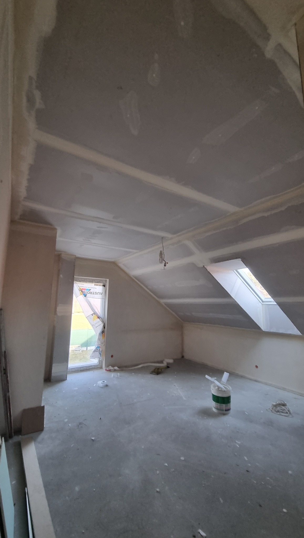 Poddasze po montażu płyt gipsowo-kartonowych, widoczne szpachlowane łączenia. Okno dachowe i okno pionowe z folią ochronną. Na podłodze resztki materiałów.