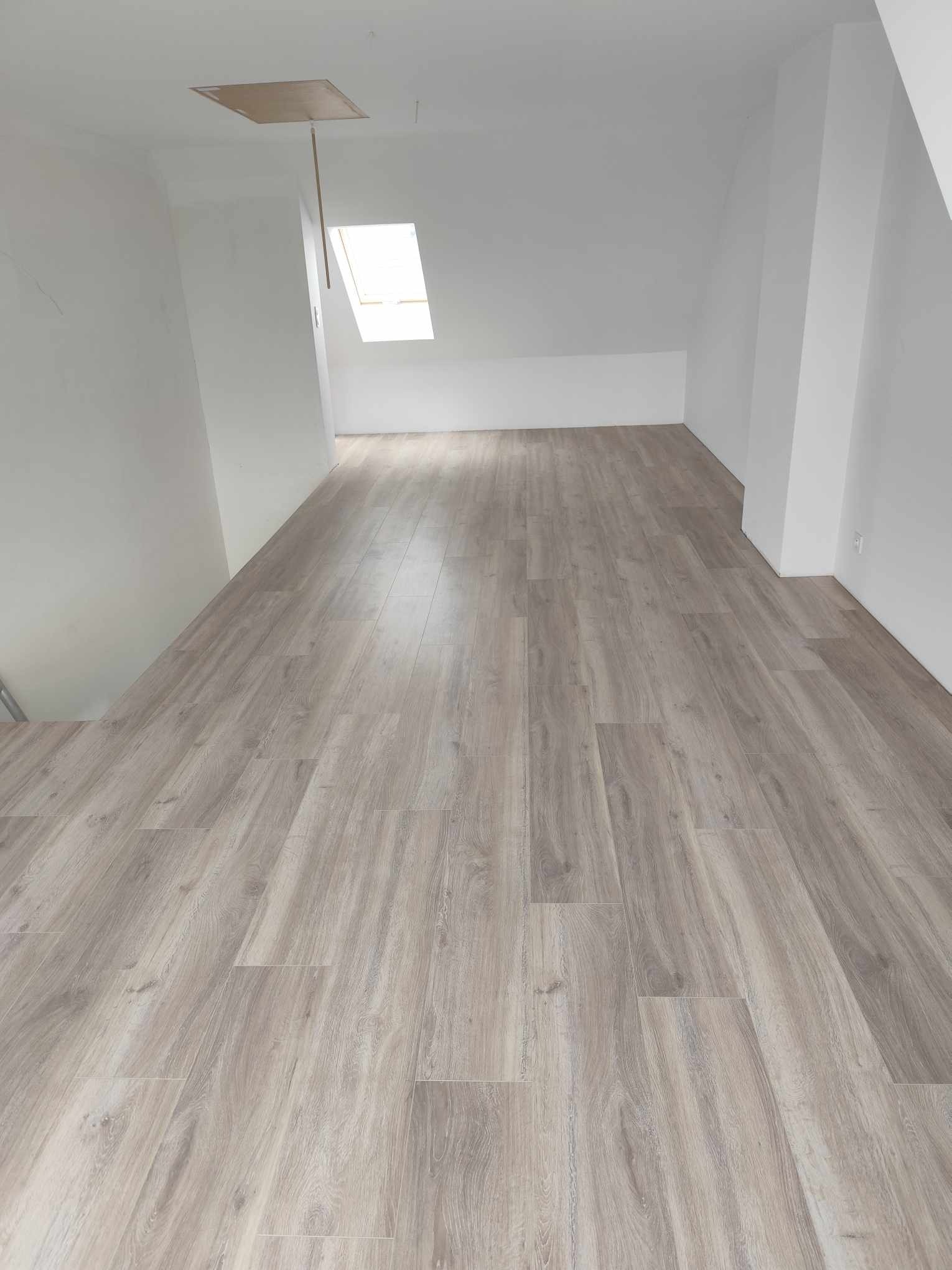 Jasne panele podłogowe w odcieniach szarości, ułożone w nowym pokoju na poddaszu z oknem dachowym i białymi ścianami. Widoczna klapa na suficie.