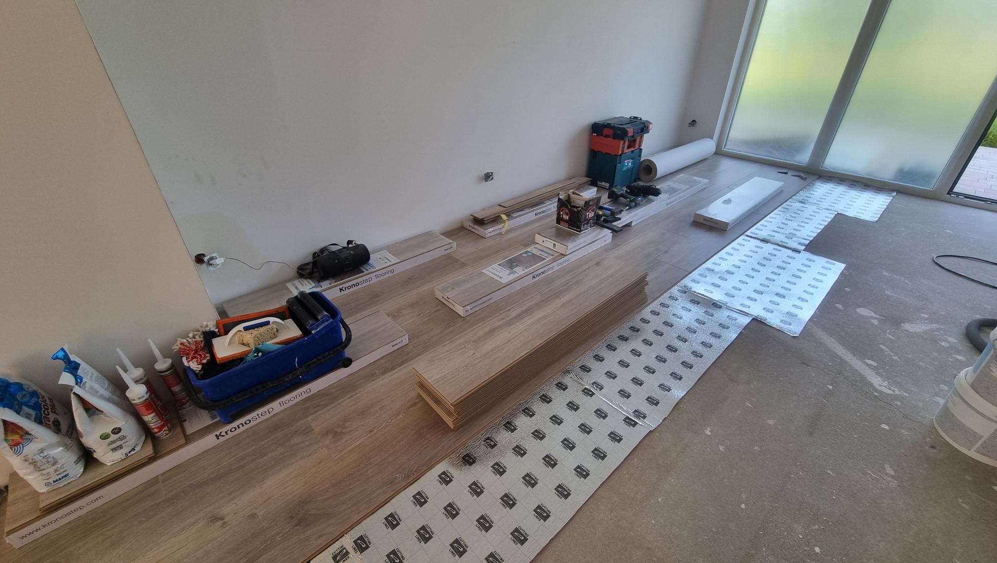 Podkład pod panele ułożony na podłodze, przygotowany do montażu paneli podłogowych. Widoczne narzędzia i materiały potrzebne do pracy. Częściowo ułożone panele podłogowe.