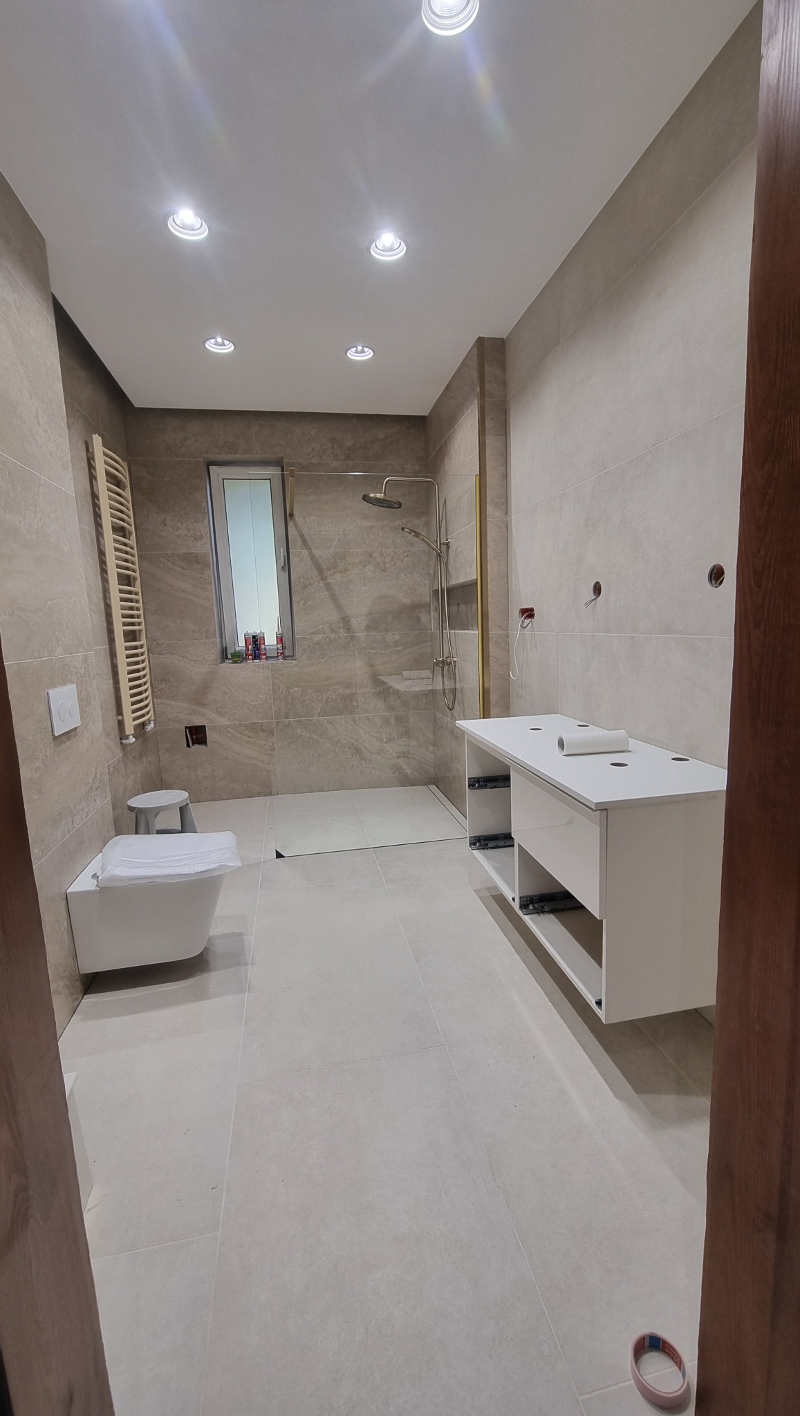 Jasna łazienka z beżowymi płytkami imitującymi kamień. Kabina prysznicowa ze złotymi detalami, biała szafka pod umywalkę i wisząca toaleta. Oświetlenie punktowe.