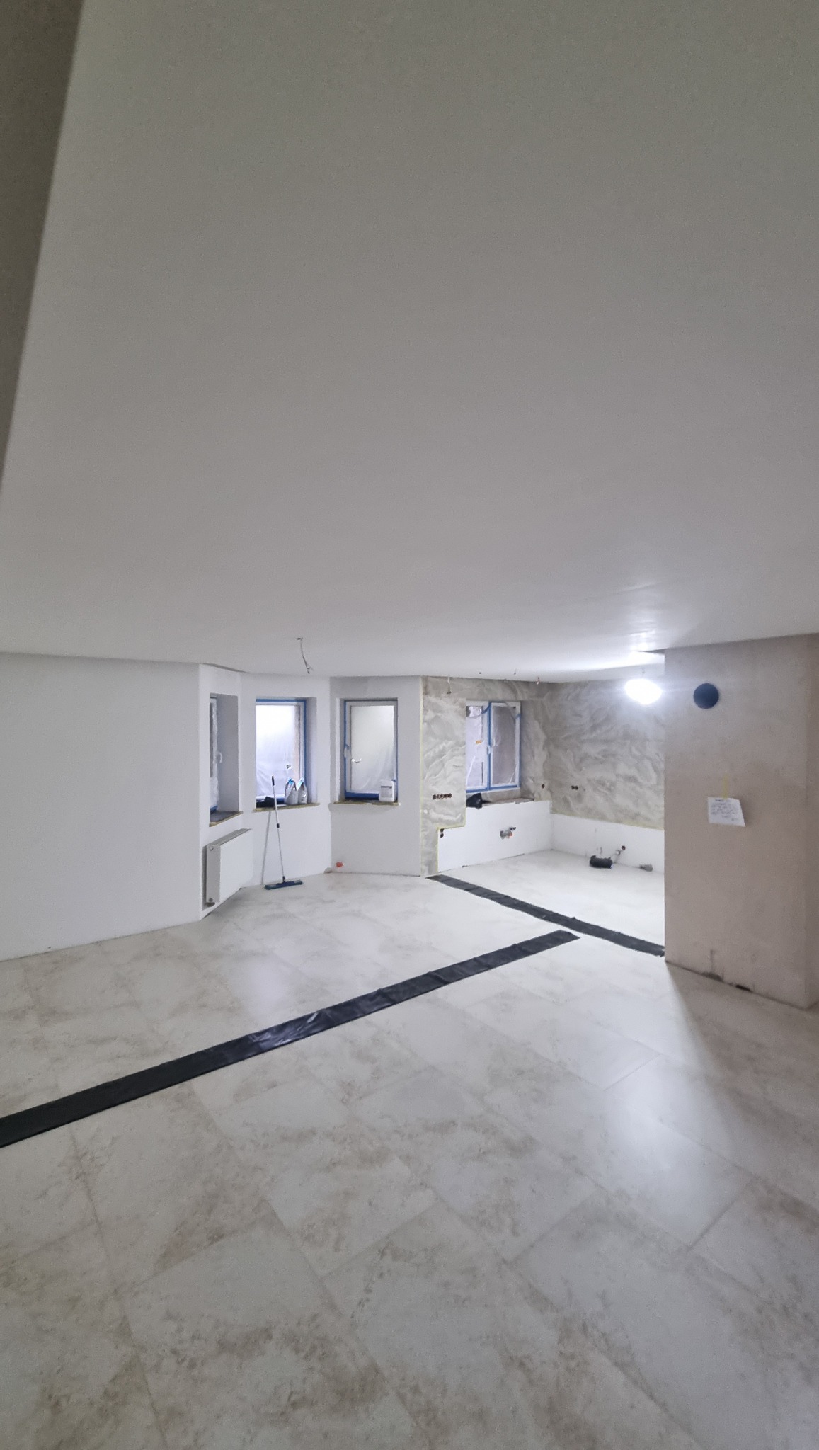 Jasne wnętrze w trakcie prac wykończeniowych: świeżo położona podłoga z płytek, częściowo pomalowane ściany, okna zabezpieczone folią malarską.