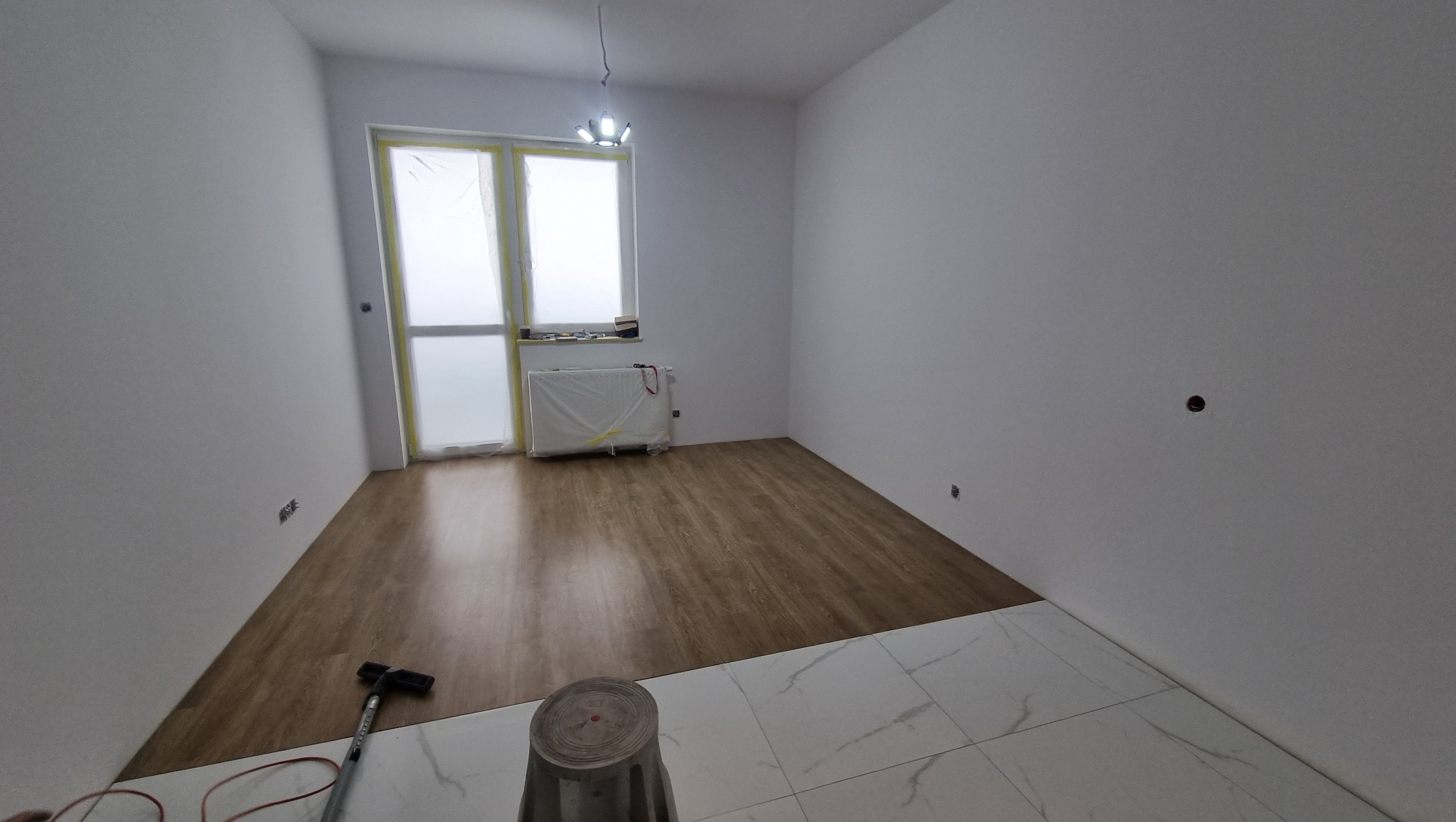 Układanie podłogi w Oławie: panele drewnopodobne zestawione z białymi płytkami. Okno zabezpieczone folią, grzejnik zakryty. Widoczne narzędzia.