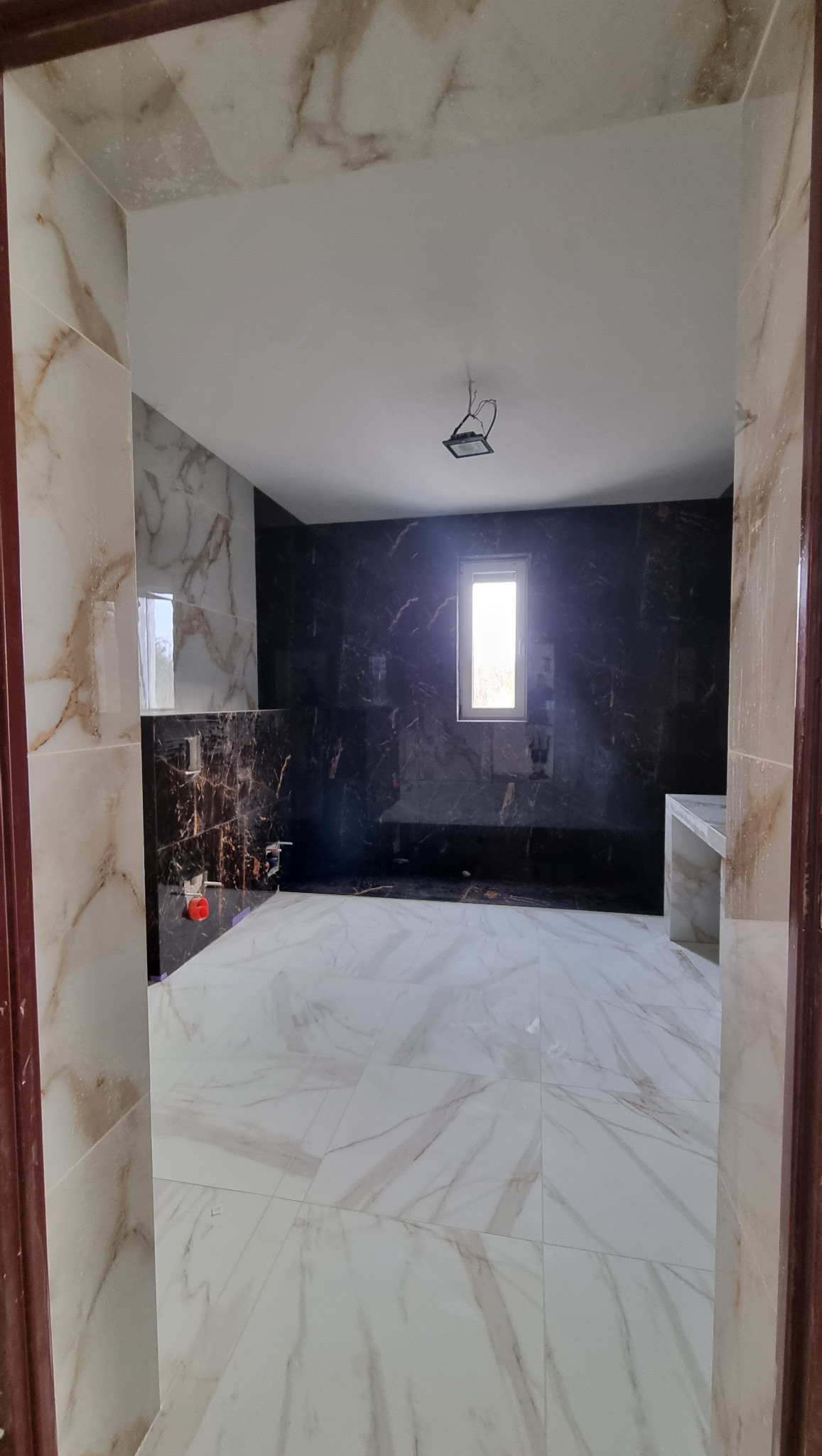 Nowoczesna łazienka z marmurowymi płytkami w odcieniach bieli i brązu, ciemna ściana z oknem, widok zza framugi drzwi. Minimalistyczny design.