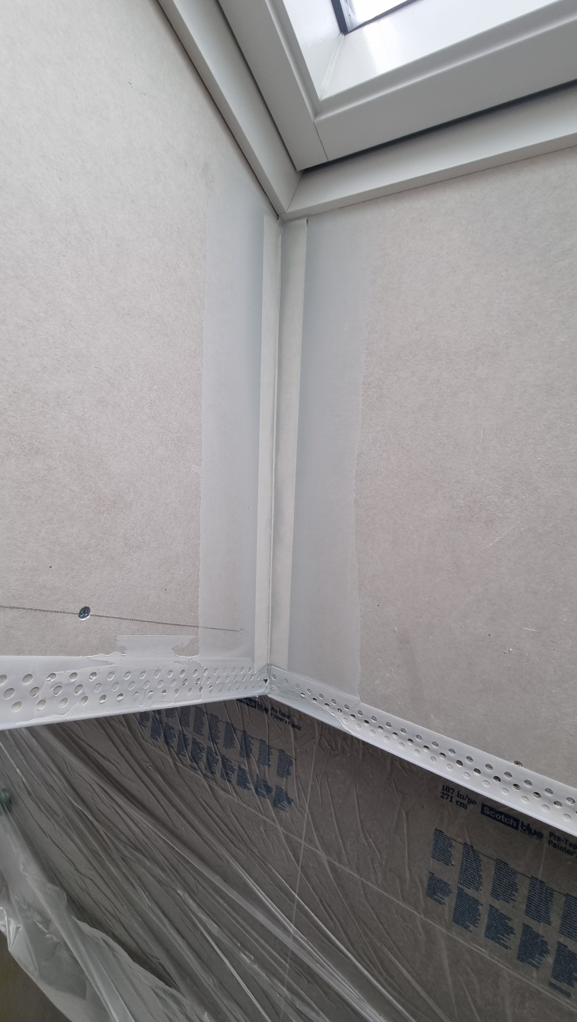 Narożnik ściany z oknem dachowym, zabezpieczony folią malarską, z zamontowanymi profilami narożnikowymi perforowanymi.