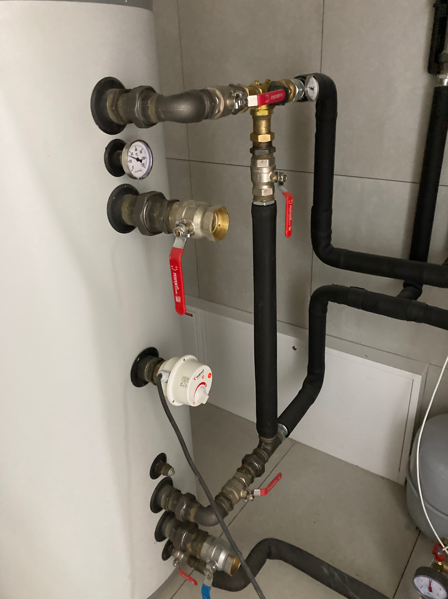 Instalacja grzewcza z zaworami, manometrem i rurami w izolacji, zamontowana przy białym zbiorniku. Widoczne elementy miedziane i czarne rury.