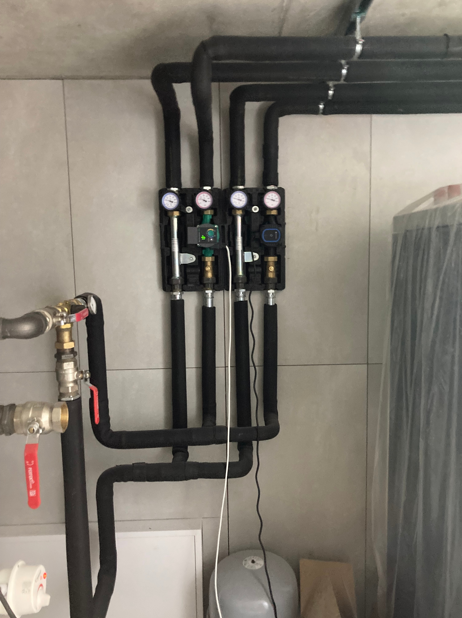Czarny rozdzielacz z manometrami i rurami w izolacji, zamontowany na jasnej ścianie. Widoczne zawory i elementy instalacji grzewczej. Część zbiornika po prawej stronie.
