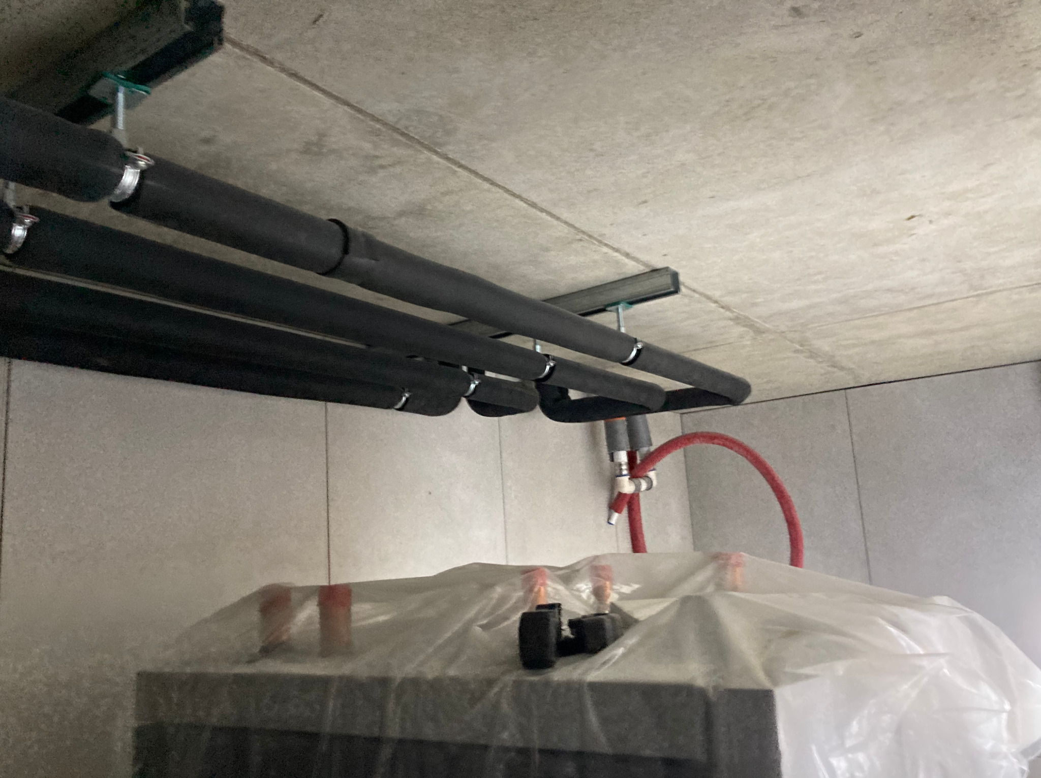 Cztery czarne, zaizolowane rury pod sufitem, zamocowane metalowymi uchwytami. Czerwona rura i elementy instalacji za folią ochronną. Szare ściany i sufit.