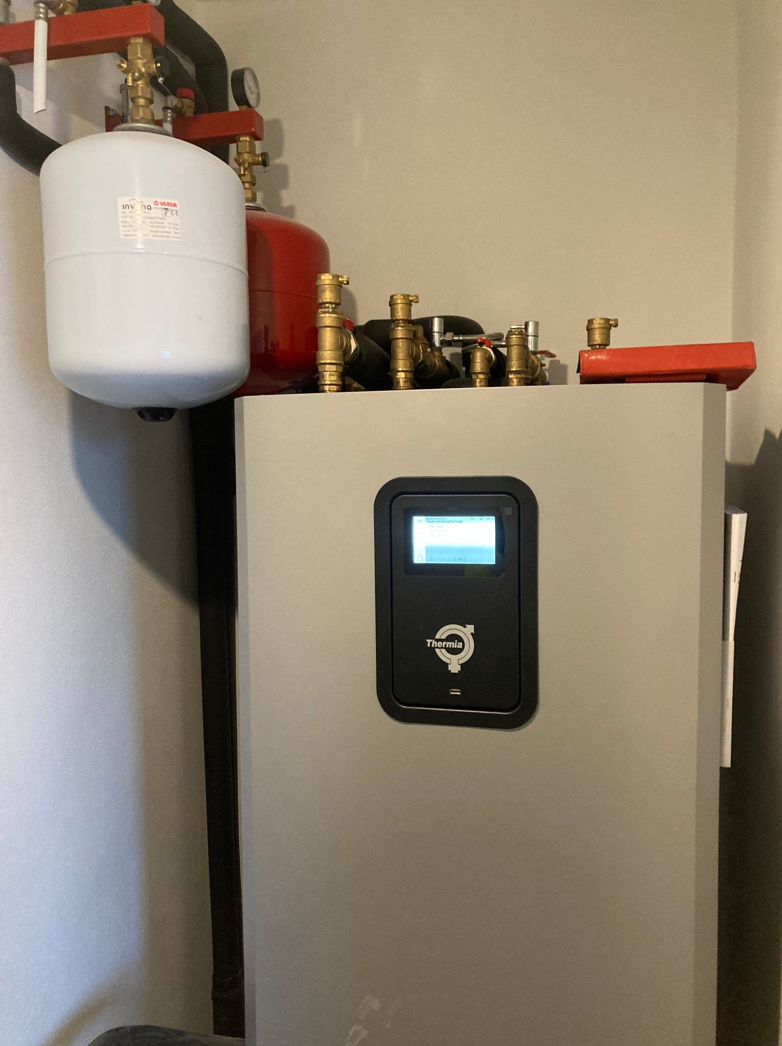 Szara pompa ciepła Thermia z wyświetlaczem, widoczne zbiorniki wyrównawcze i instalacja hydrauliczna. Ujęcie z dołu, oświetlenie naturalne.