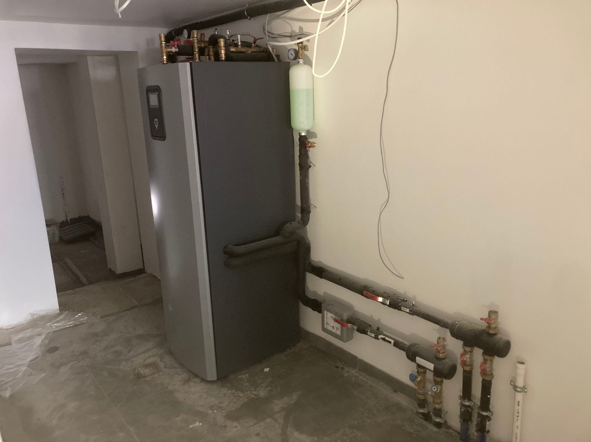 Szara pompa ciepła Thermia z podłączonymi rurami w surowym wnętrzu. Widoczne elementy instalacji i manometr. Ujęcie z boku, z bliska.