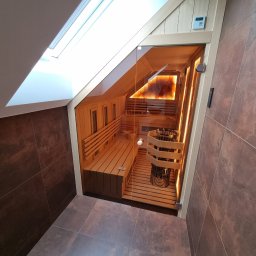 JC Dom Jan Chodacki - Sauna w poddaszu z drewnianymi ławkami i piecem, oświetlona lampami solnymi. Obudowa z ciemnych płytek, widoczne okno dachowe. Całość utrzymana w ciepłej tonacji.