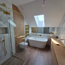 Nowoczesna łazienka na poddaszu z drewnianą podłogą i szafką, białą wanną wolnostojącą i złotymi akcentami w armaturze oraz kabinie prysznicowej.