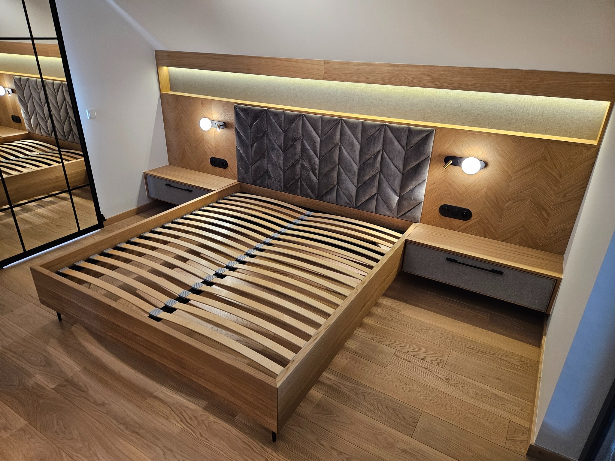 Nowoczesna sypialnia z drewnianym łóżkiem, tapicerowanym zagłówkiem w geometryczny wzór i oświetleniem LED. Widok z perspektywy podłogi podkreśla dębowy parkiet.