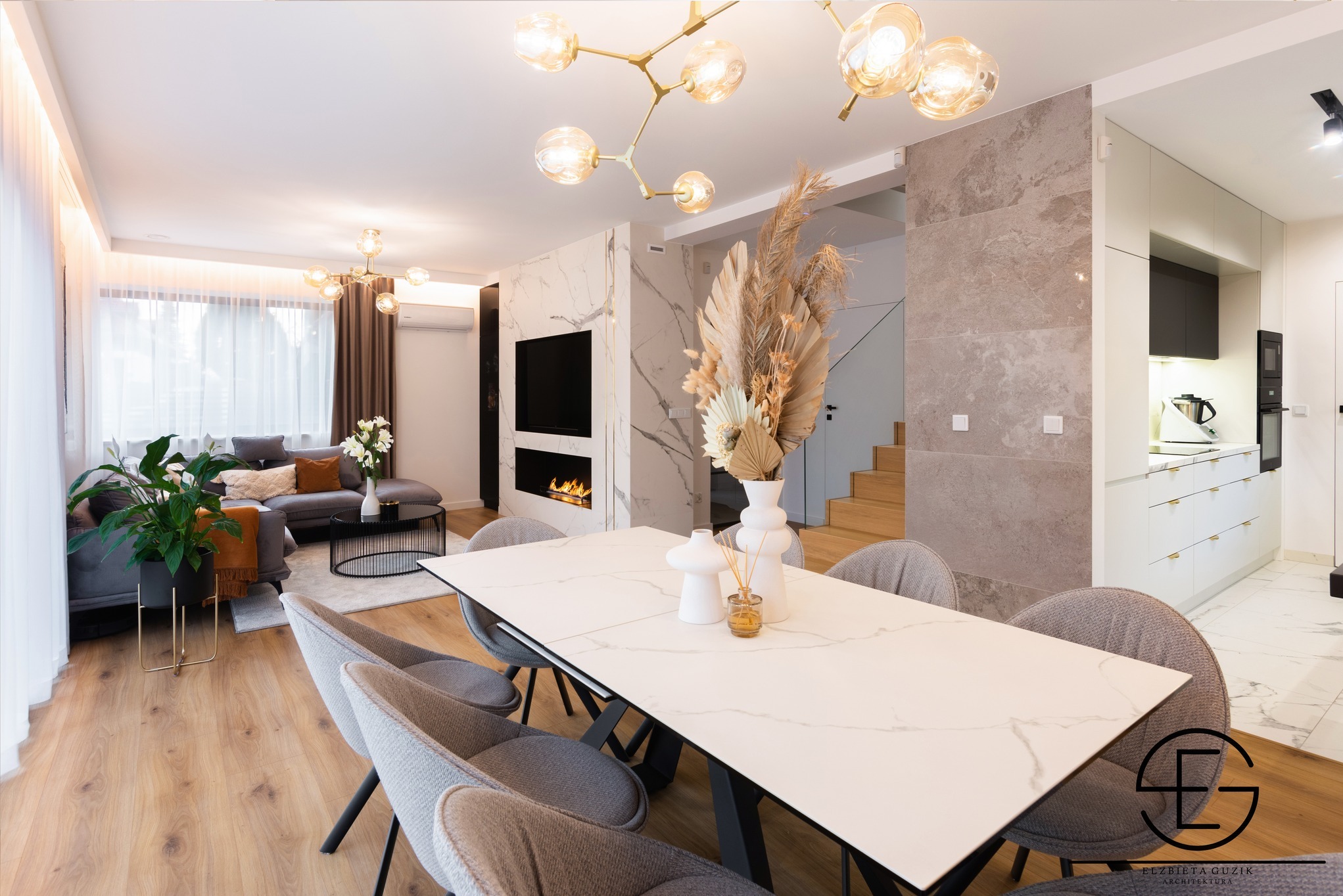 Elegancja w detalu: Jasne, przestronne wnętrze z marmurowymi akcentami, nowoczesnym oświetleniem i stylowym stołem jadalnym z szarymi krzesłami. Widoczny salon i kuchnia.