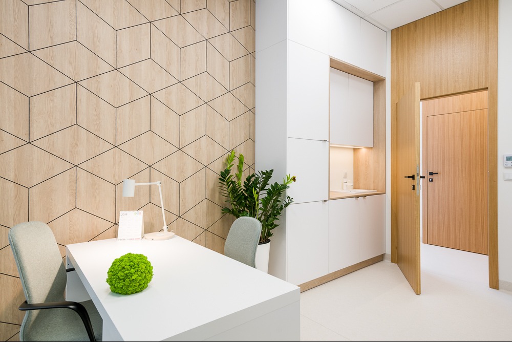 Nowoczesne biuro: geometryczna ściana, białe meble, drewniane akcenty i roślina doniczkowa na biurku, lampa biurkowa. Otwarta drewniana  drzwi w tle.