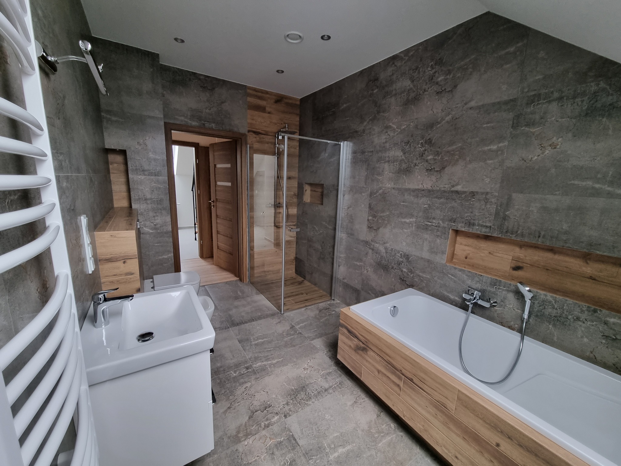 Nowoczesna łazienka z szarymi płytkami imitującymi kamień, drewnianymi akcentami i białą armaturą. Kabina prysznicowa ze szkła, grzejnik drabinkowy.