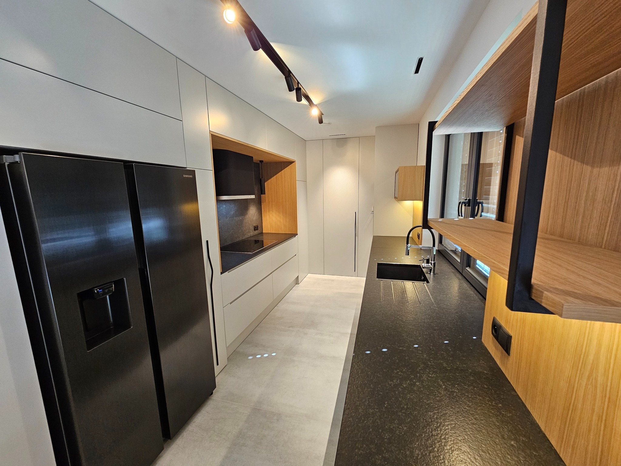 Nowoczesna kuchnia z czarną lodówką Samsung, jasnymi szafkami i drewnianymi akcentami. Oświetlenie szynowe podkreśla minimalistyczny design i czarne blaty.