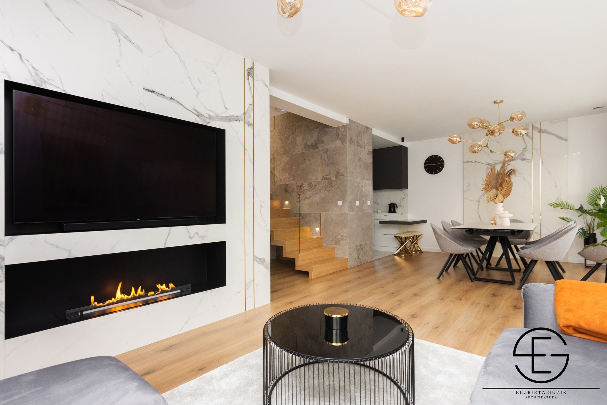 Elegancki salon z kominkiem i telewizorem w zabudowie z marmuru, schody z drewnianymi stopniami. Nowoczesne wykończenie wnętrza z dbałością o detale.