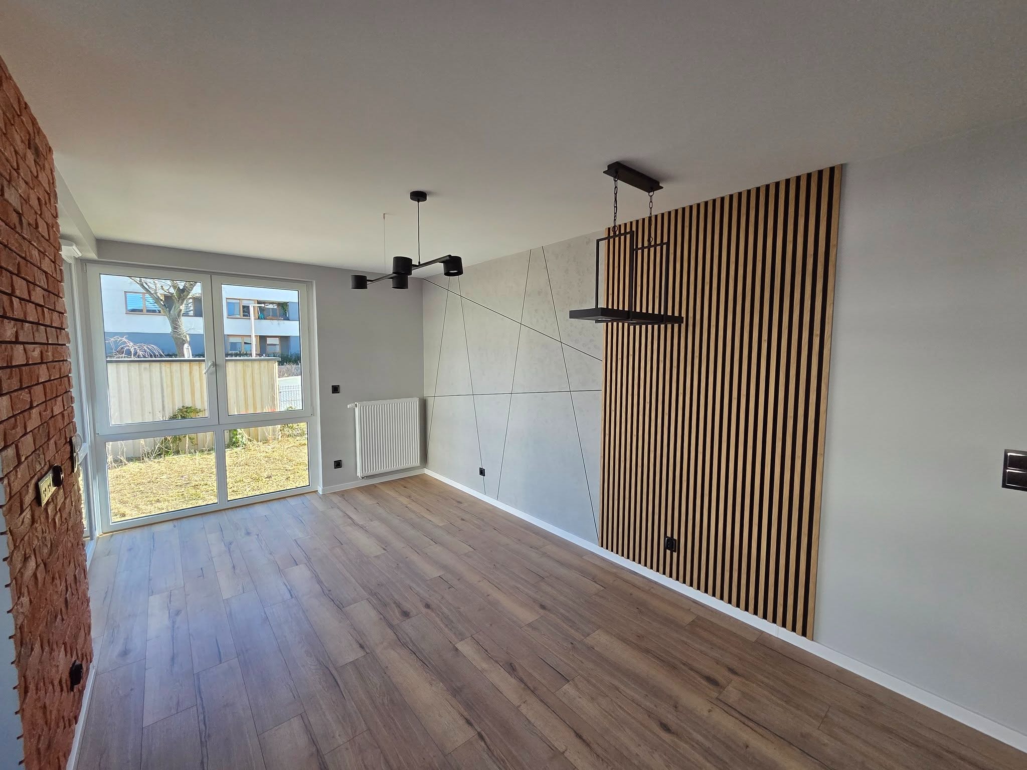 Wykończone wnętrze pokoju z drewnianą podłogą, panelami ściennymi i nowoczesnym oświetleniem. Widok na okno i fragment ceglanej ściany.