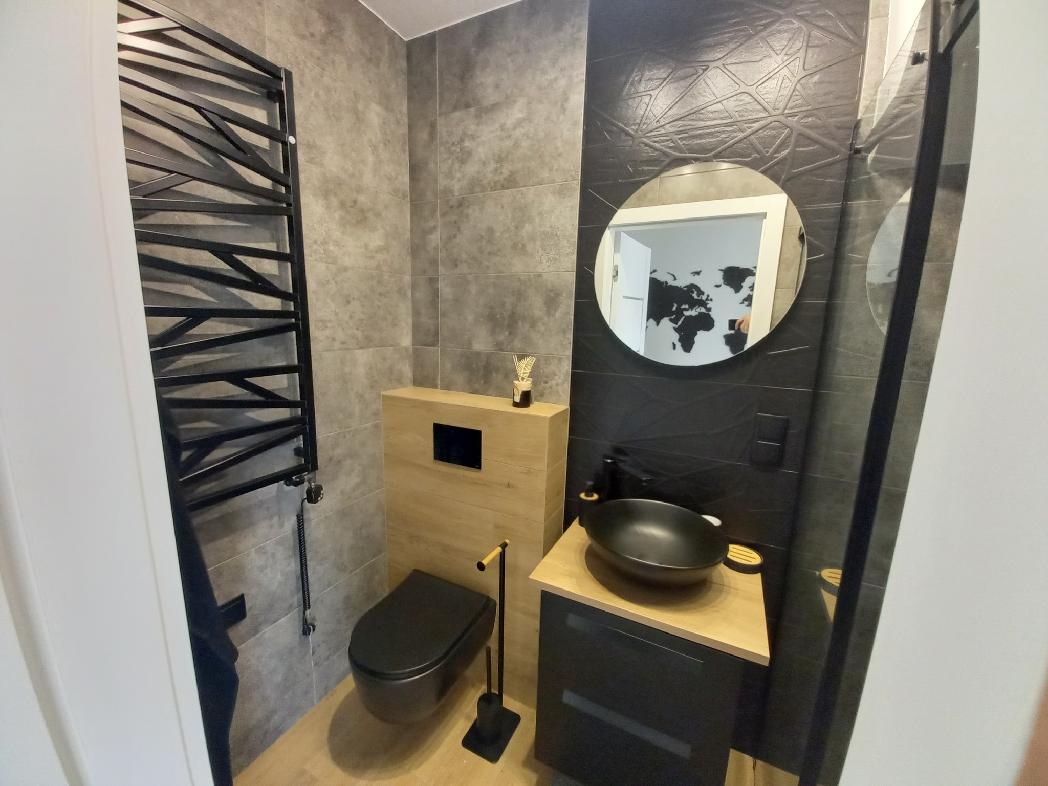 Nowoczesna łazienka z czarną armaturą: umywalka nablatowa, wisząca toaleta, grzejnik drabinkowy. Szare płytki i drewnopodobne akcenty tworzą elegancką przestrzeń.