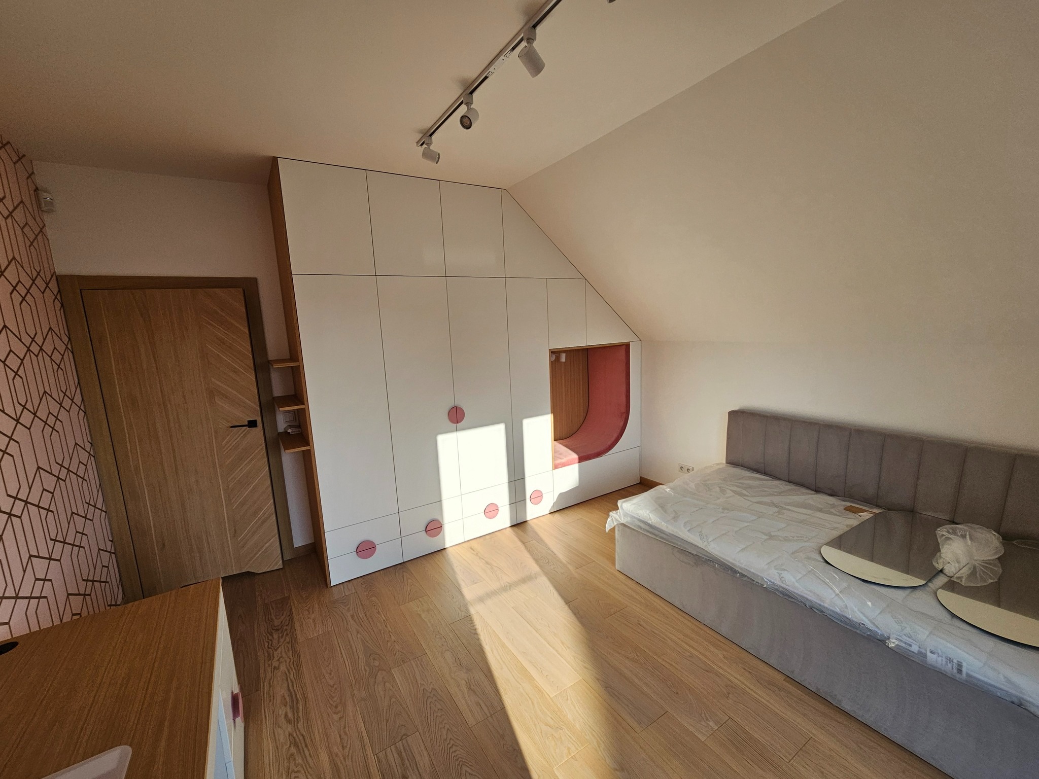 Pokój na poddaszu z zabudową meblową: białe szafy z różowymi akcentami, wnęka z siedziskiem, drewniana podłoga i drzwi. Nowoczesne oświetlenie punktowe.