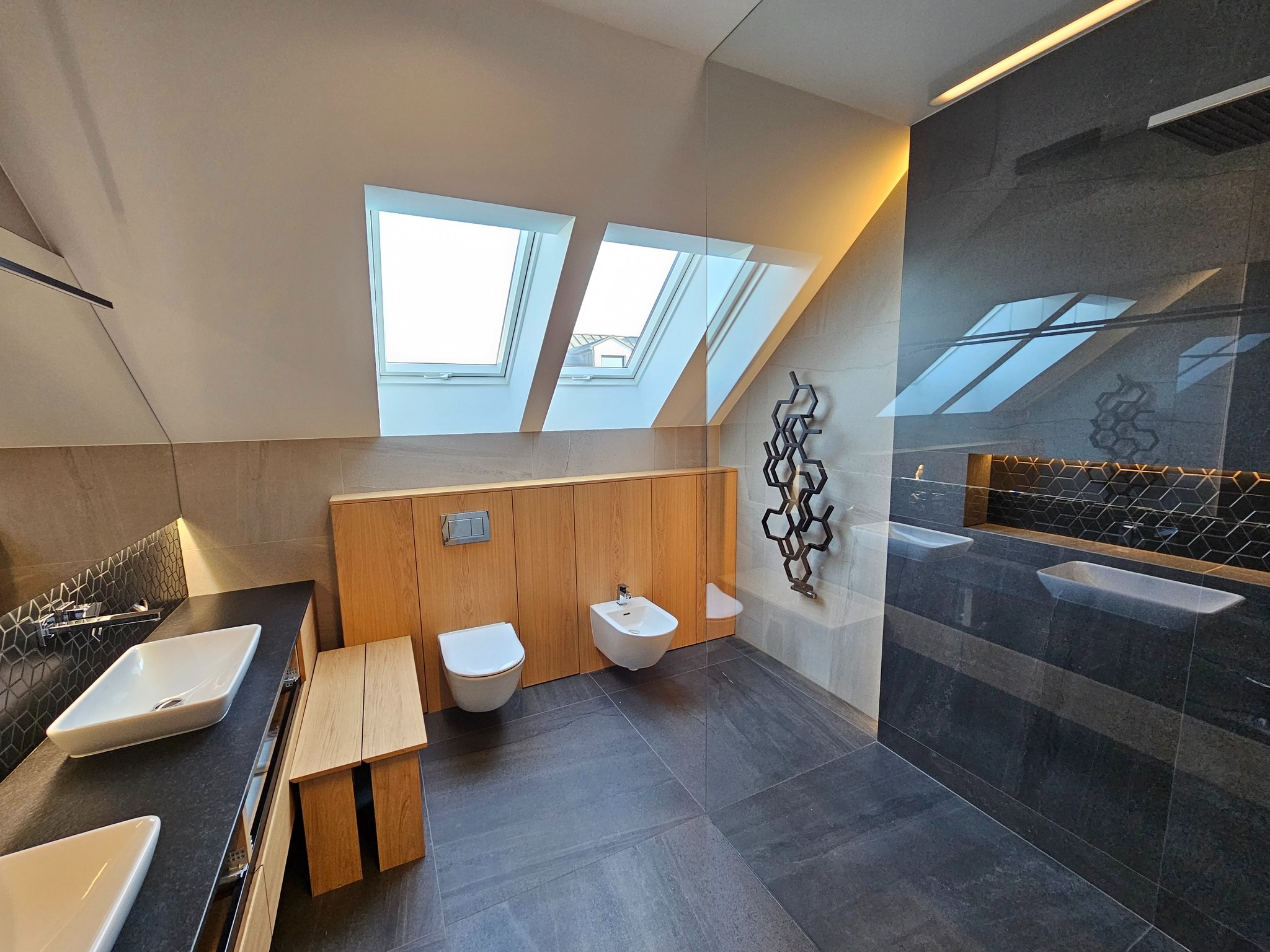 Nowoczesna łazienka na poddaszu z oknami dachowymi, drewnianą zabudową, czarną armaturą i płytkami w odcieniach szarości. Minimalistyczny design z akcentami drewna i geometrycznymi dekoracjami.