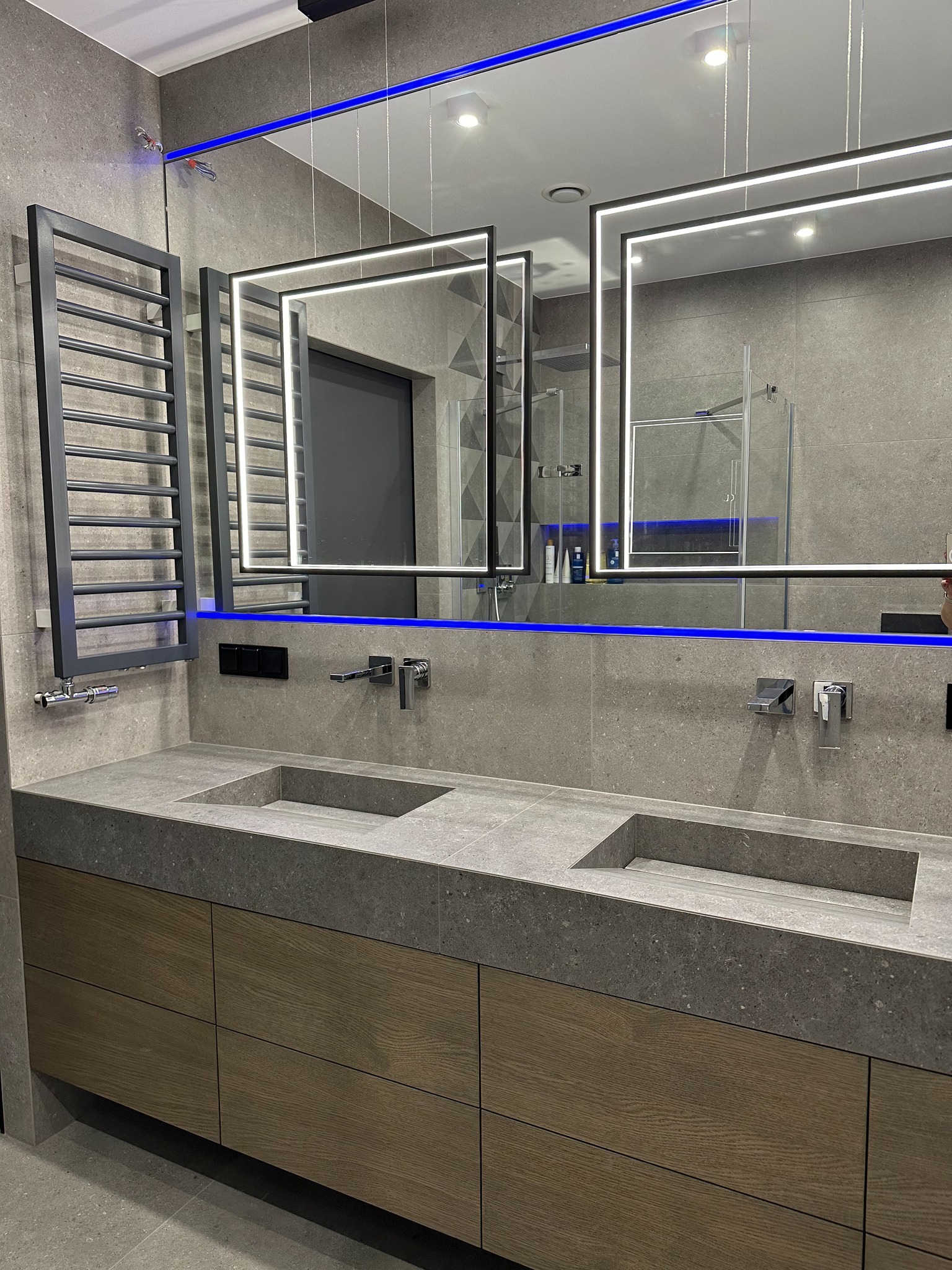 Nowoczesna łazienka z podwójną umywalką z szarego kamienia, podświetlanymi lustrami i grzejnikiem drabinkowym. Minimalistyczny design z akcentami niebieskiego światła.