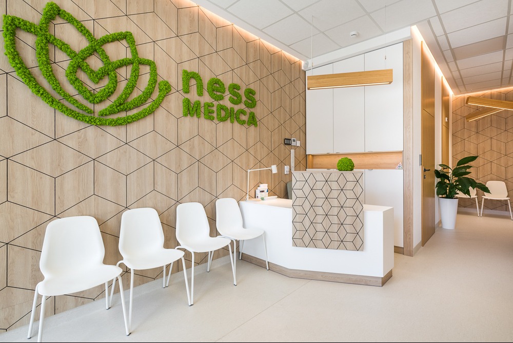 Nowoczesna recepcja z drewnianą ścianą w geometryczne wzory, zielonym logo z mchu i białymi krzesłami w jasnym, przestronnym wnętrzu.
