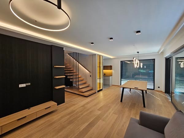 Nowoczesne wnętrze z drewnianą podłogą, czarną ścianą TV, schodami i oświetleniem LED. Minimalistyczny design z elementami drewna i czerni.