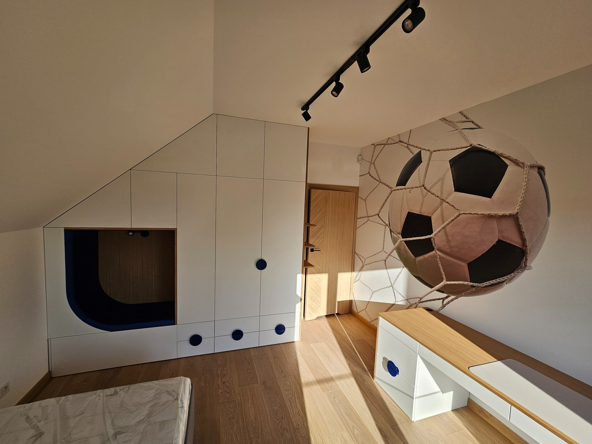Pokój dziecięcy na poddaszu: biała zabudowa meblowa z wnęką do spania, drewniana podłoga, dekoracja ściany z motywem piłki nożnej. Nowoczesne oświetlenie punktowe.