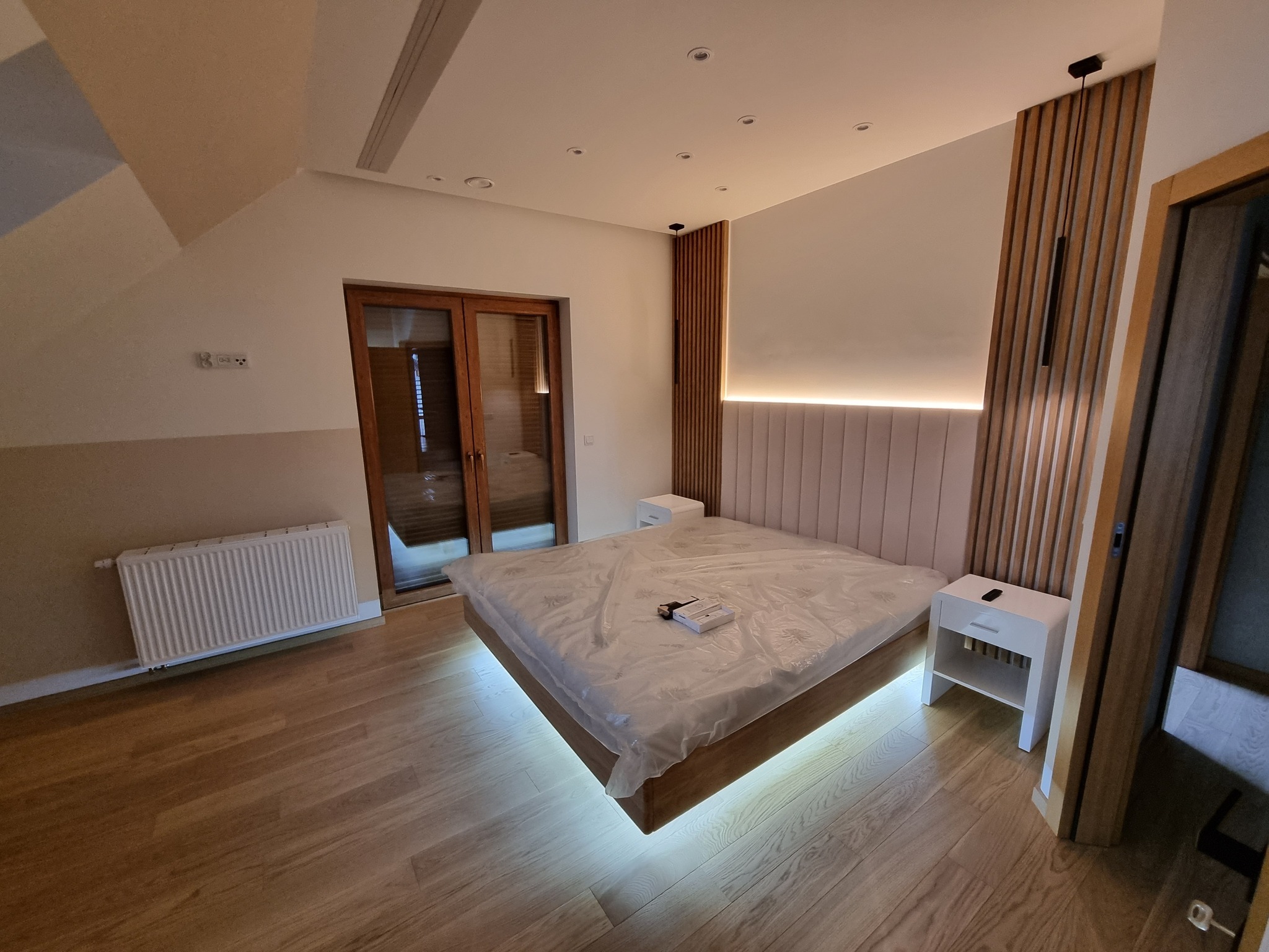 Nowoczesna sypialnia na poddaszu z drewnianą podłogą i podświetlanym łóżkiem, częściowo jeszcze zabezpieczonym folią, po remoncie. Minimalistyczny design.