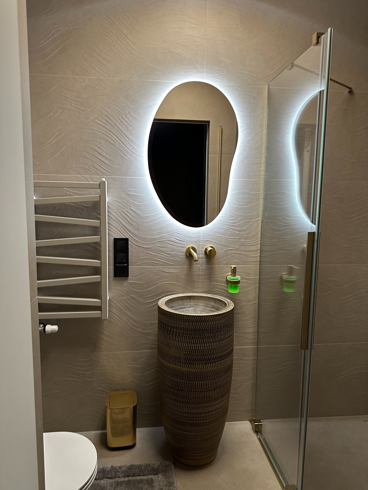 Nowoczesna łazienka z umywalką w formie wazonu, podświetlanym lustrem o nieregularnym kształcie i kabiną prysznicową ze złotymi detalami. Na ścianach płytki z motywem fal.
