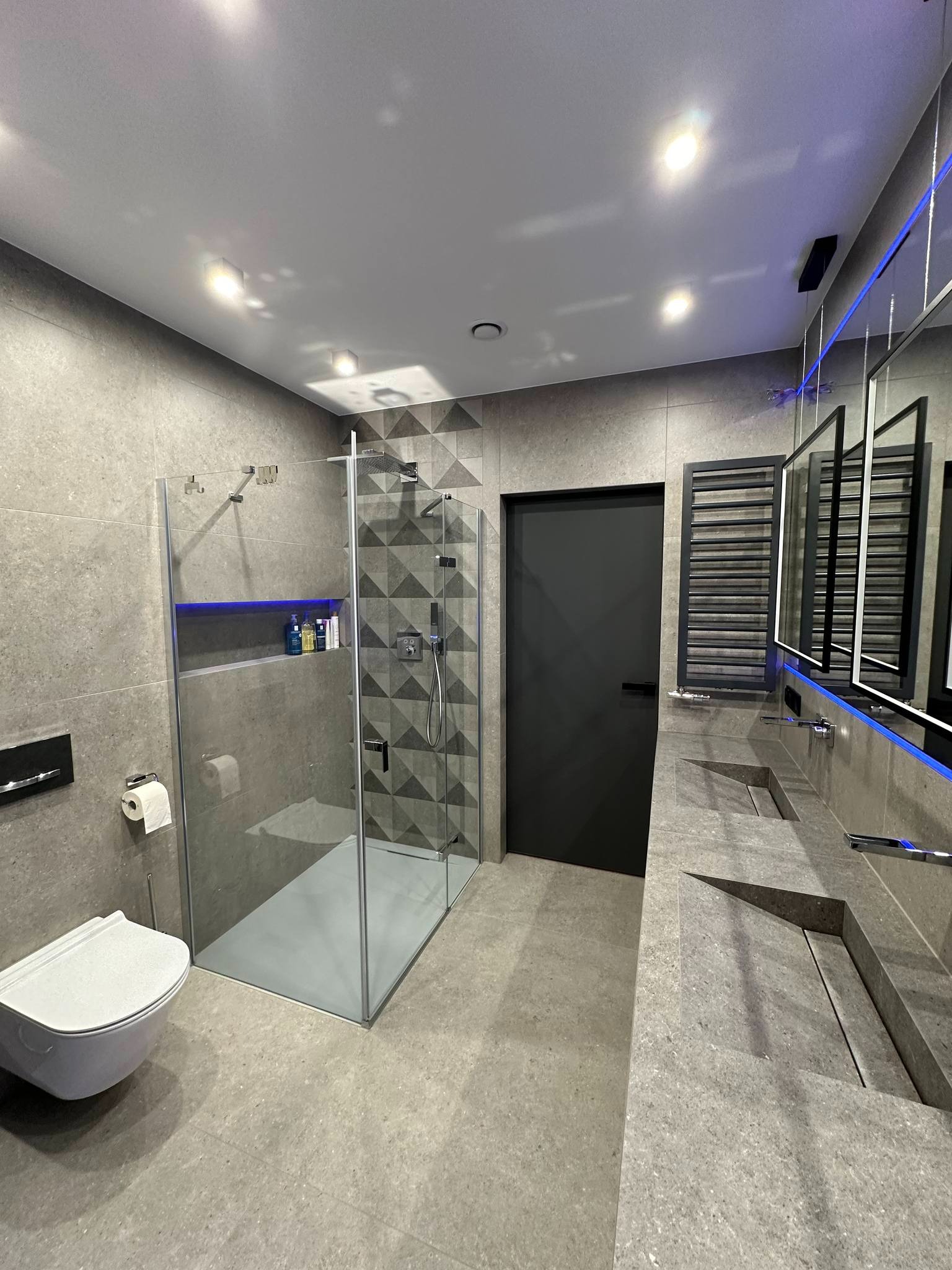 Nowoczesna łazienka w szarościach: kabina prysznicowa ze wzorem geometrycznym, podwieszana toaleta, umywalka wpuszczana w blat, lustra z podświetleniem LED.